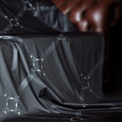 Black printed water proof and lube proof throw with a square design of black leather bondage straps with silver metal connectors shown draped over a brown leather Chesterfield sofa with part of the arm showing