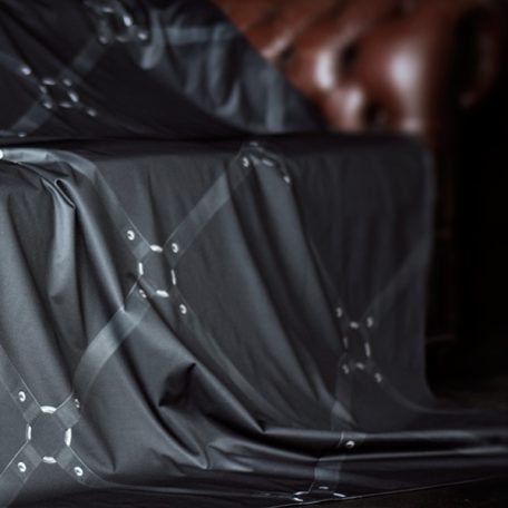 Black printed water proof and lube proof throw with a square design of black leather bondage straps with silver metal connectors shown draped over a brown leather Chesterfield sofa with part of the arm showing